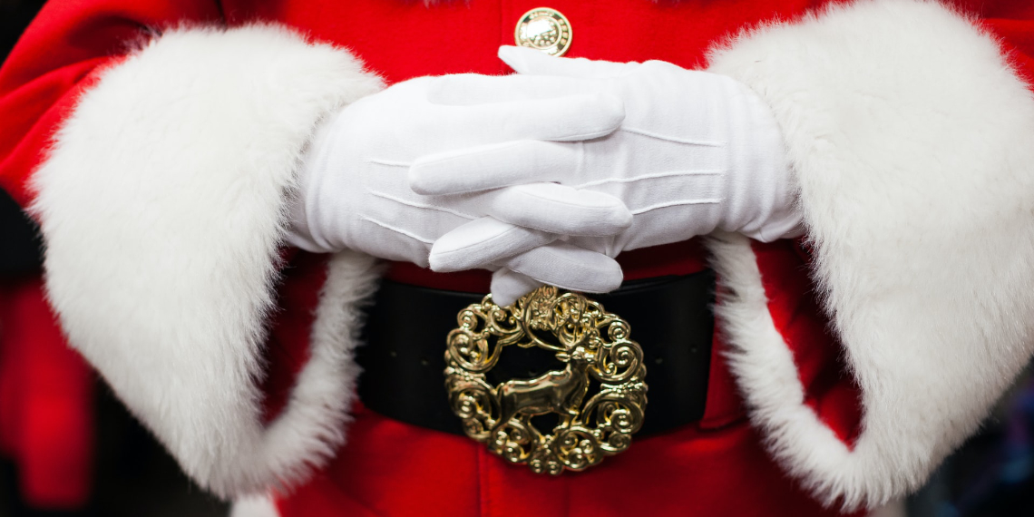 Close-up of Santa Claus white gloved hands over a red suit and black belt with a stag motif on the brass belt buckle